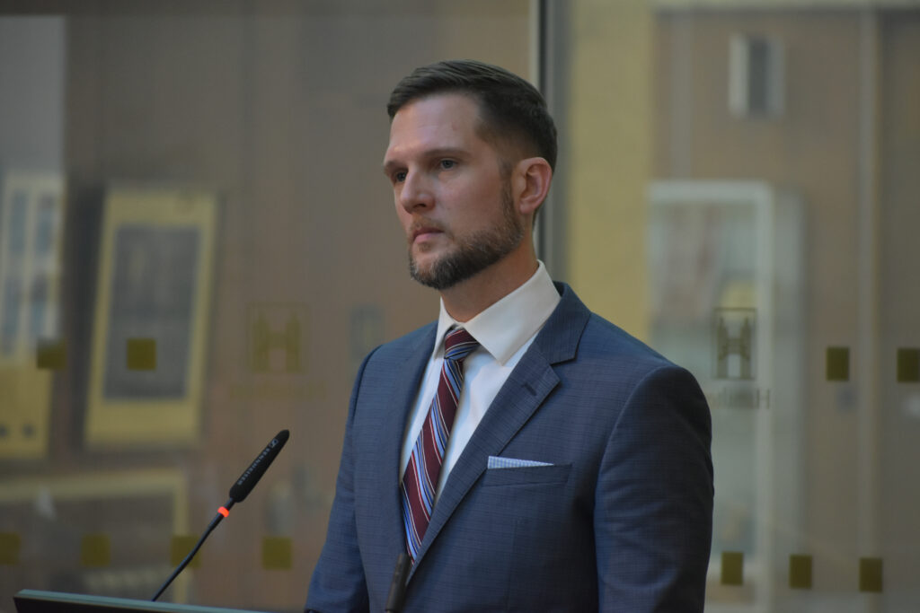 A man, Keanin Loomis, wearing a suit and tie, standing at a podium.