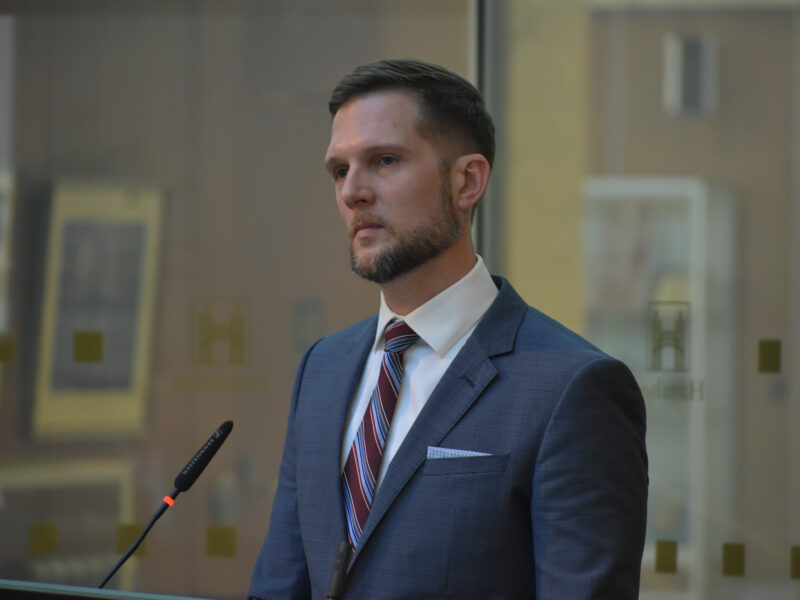 A man, Keanin Loomis, wearing a suit and tie, standing at a podium.