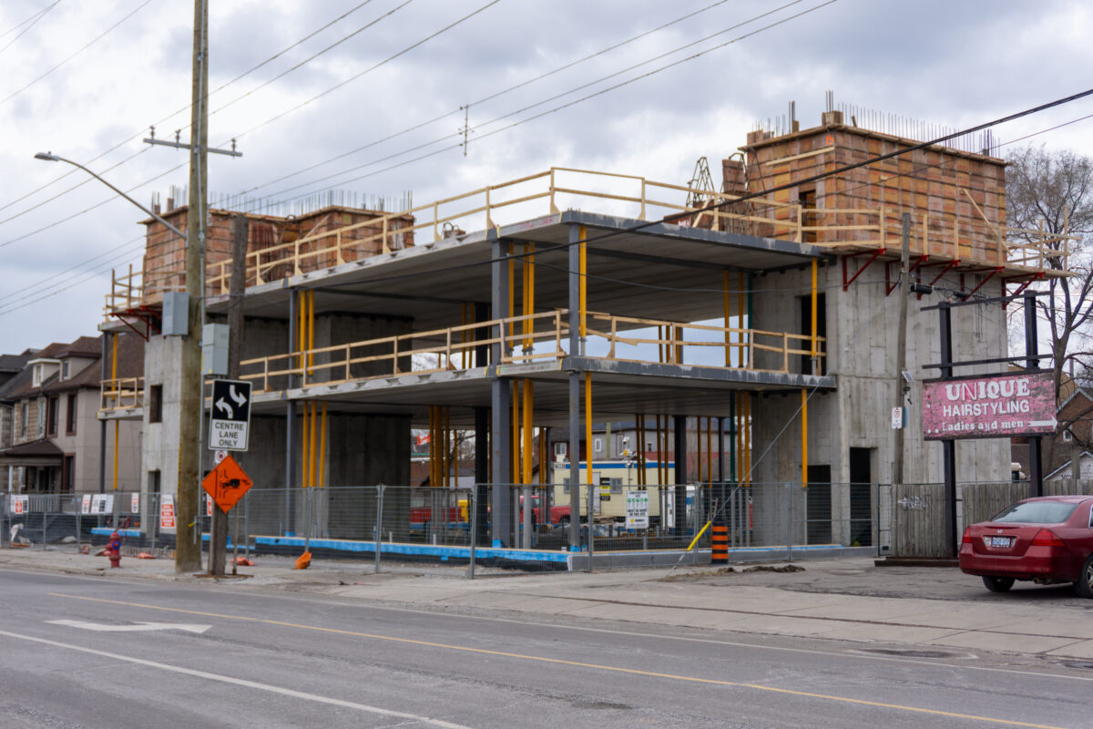A construction site for a multi-story building. The concrete foundation and first two floors of a steel-and-concrete frame are visible, supported by a dense forest of yellow metal shoring posts. The site is enclosed by a chain-link fence next to a city sidewalk. Image 7: King