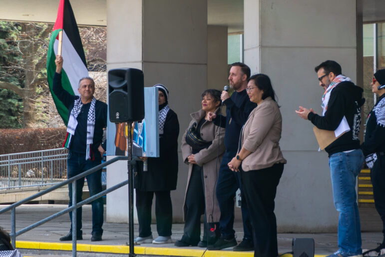A group of people stand under the overhang of City Hall. A man speaks into a microphone while others, including city councillors, listen. Several individuals are wearing Keffiyeh scarves.