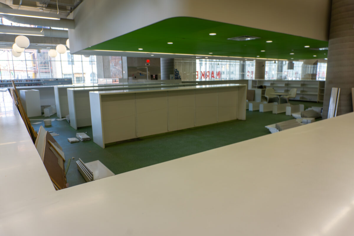 Interior of a library showing rows of empty white bookshelves and green carpeting. The area has been cleared of books and appears to be in the process of being dismantled.