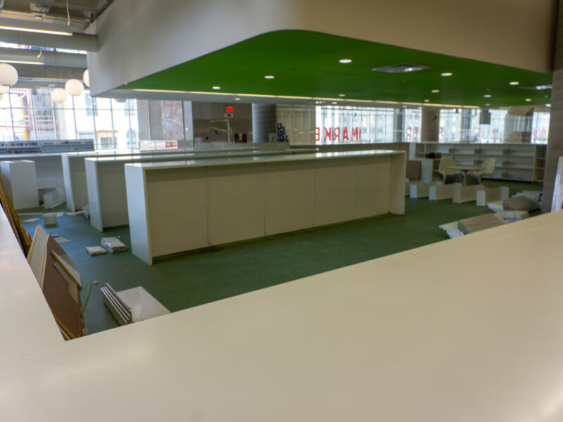 Interior of a library showing rows of empty white bookshelves and green carpeting. The area has been cleared of books and appears to be in the process of being dismantled.