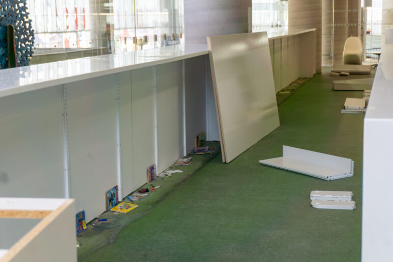 Close-up of green library carpeting where shelves used to stand. A few thin children's books and dust are left on the floor near a white wall.