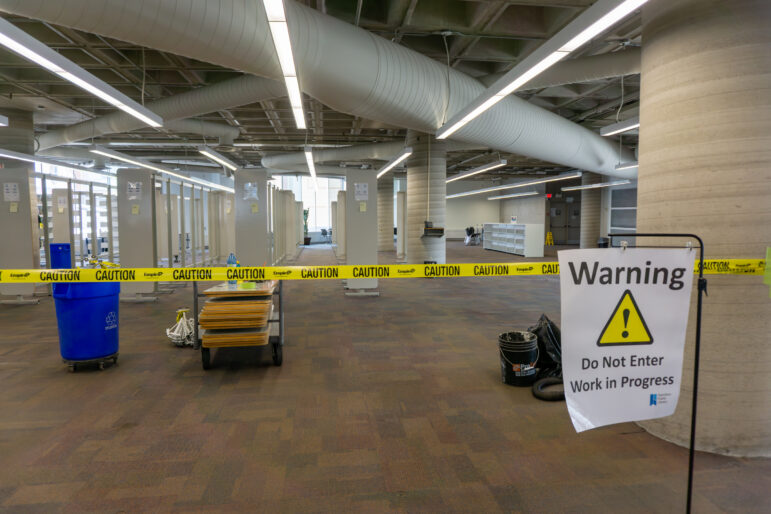 A large open library space with grey carpeting. Yellow caution tape and a sign stating "Warning: Do Not Enter, Work in Progress" block the foreground.