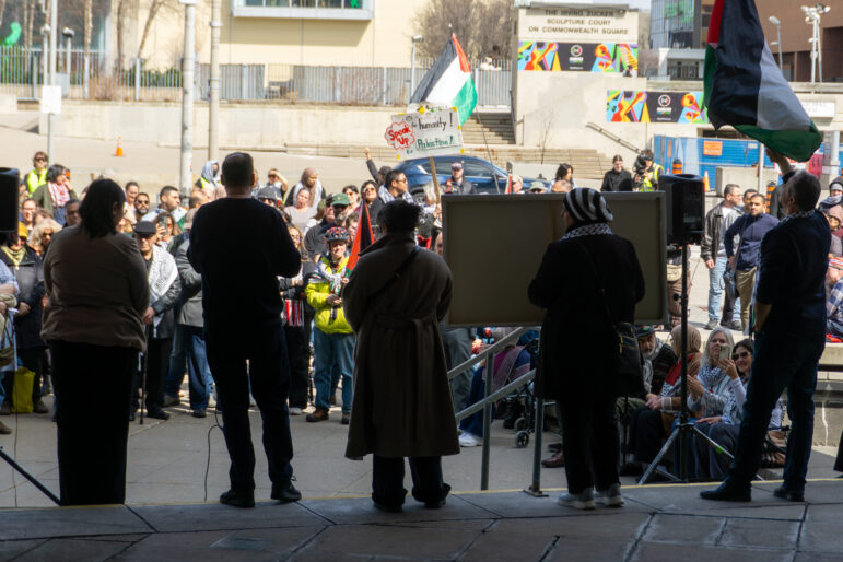 A view from behind several speakers on a raised platform looking out at a large crowd gathered in front of Hamilton City Hall. One attendee holds a Palestinian flag.