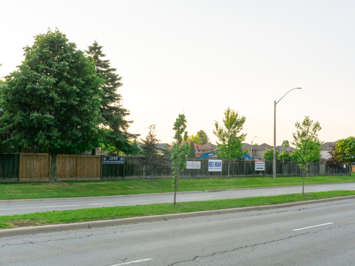 A large "Coming Soon" development sign for Kiwanis Homes stands in a grassy field near a sidewalk. The sign features an architectural rendering of a modern, four-story residential building with light-colored siding and large windows. The logos for Kiwanis Homes, the City of Hamilton, and the Ontario and Canada governments are prominently displayed at the bottom. In the background, a residential street with mature trees is visible under a clear blue sky.
