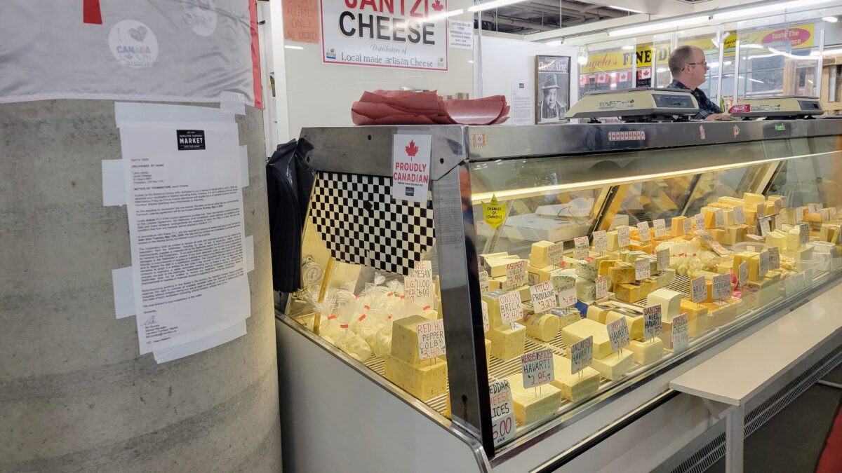 A wide shot inside the Hamilton Farmers’ Market showing the Jantzi Cheese stall. A refrigerated glass display case in the center is filled with various blocks and bags of cheese with price tags. To the left of the counter, a white paper notice is taped to a large, round concrete pillar. In the background, signs for the business and other market stalls are visible.