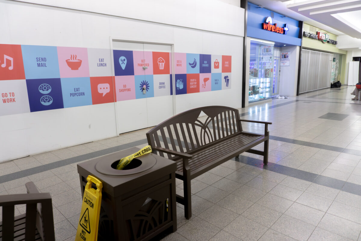 A quiet interior hallway of Jackson Square mall. In the foreground, a brown wooden bench sits next to a matching square garbage can. A strip of yellow "POLICE LINE DO NOT CROSS" tape is draped into the opening of the trash bin. A yellow "Caution Wet Floor" sign stands on the tile floor nearby. In the background, storefronts for "Wireless +" and "Pita Pit" are visible under bright overhead lighting.