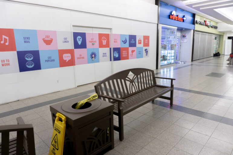 A quiet interior hallway of Jackson Square mall. In the foreground, a brown wooden bench sits next to a matching square garbage can. A strip of yellow "POLICE LINE DO NOT CROSS" tape is draped into the opening of the trash bin. A yellow "Caution Wet Floor" sign stands on the tile floor nearby. In the background, storefronts for "Wireless +" and "Pita Pit" are visible under bright overhead lighting.