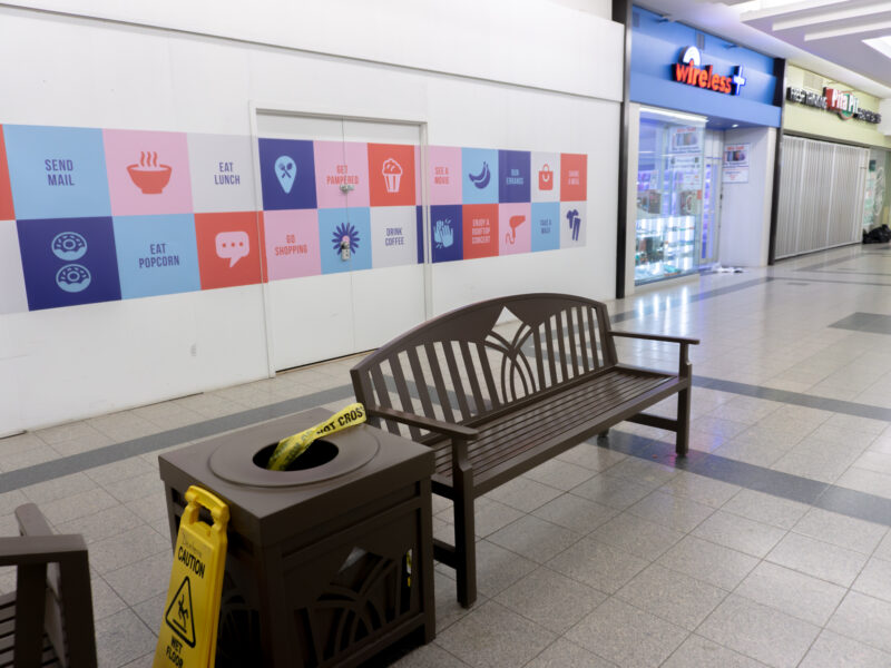 A quiet interior hallway of Jackson Square mall. In the foreground, a brown wooden bench sits next to a matching square garbage can. A strip of yellow "POLICE LINE DO NOT CROSS" tape is draped into the opening of the trash bin. A yellow "Caution Wet Floor" sign stands on the tile floor nearby. In the background, storefronts for "Wireless +" and "Pita Pit" are visible under bright overhead lighting.