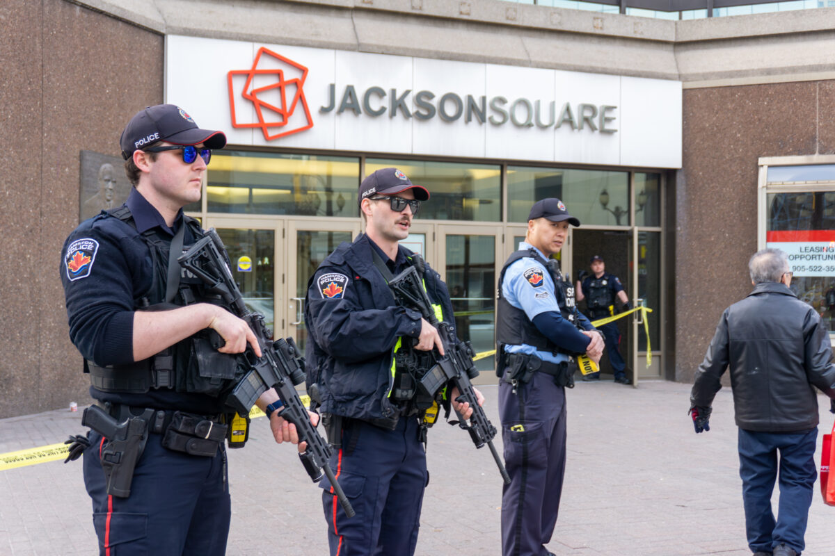 Three Hamilton Police officers stand on the sidewalk in front of the glass doors of Jackson Square. The two officers in the foreground are wearing tactical vests and holding rifles. Yellow police tape is visible behind them, cordoning off the entrance. The mall's red and grey "Jackson Square" logo is mounted on the wall above the doors.