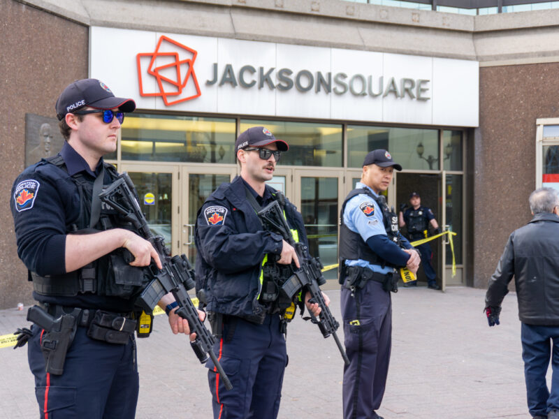 Three Hamilton Police officers stand on the sidewalk in front of the glass doors of Jackson Square. The two officers in the foreground are wearing tactical vests and holding rifles. Yellow police tape is visible behind them, cordoning off the entrance. The mall's red and grey "Jackson Square" logo is mounted on the wall above the doors.