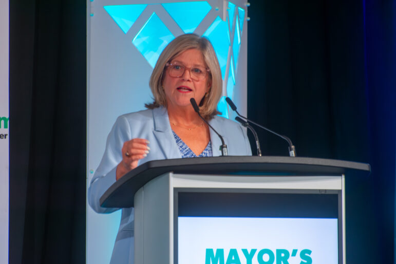A close-up shot of Mayor Andrea Horwath speaking at a podium. She is wearing glasses and a light blue blazer over a blue and white patterned shirt. The podium features a digital screen with the words "MAYOR'S BREAKFAST." Behind her is a white decorative pillar illuminated with blue geometric lights