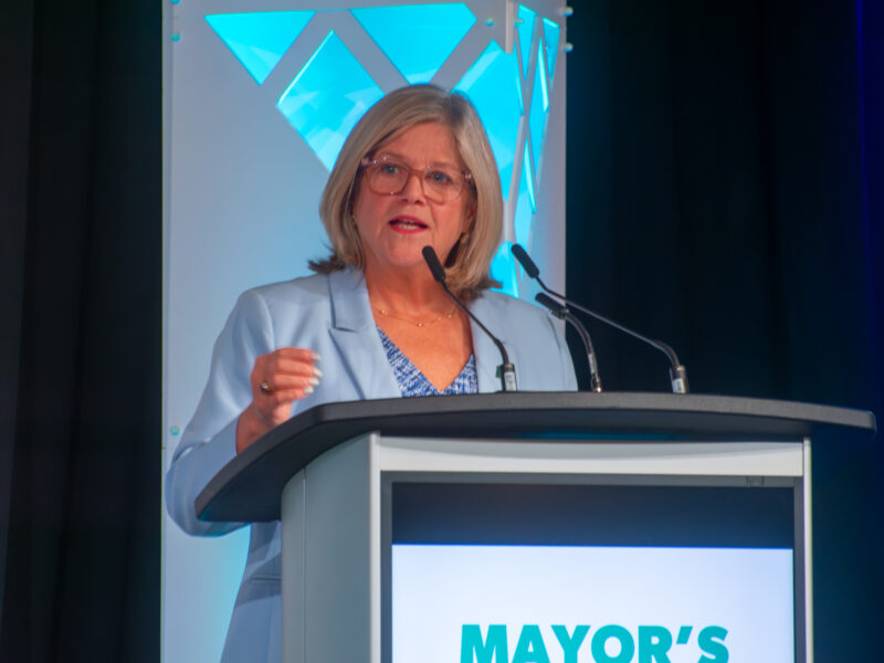 A close-up shot of Mayor Andrea Horwath speaking at a podium. She is wearing glasses and a light blue blazer over a blue and white patterned shirt. The podium features a digital screen with the words "MAYOR'S BREAKFAST." Behind her is a white decorative pillar illuminated with blue geometric lights