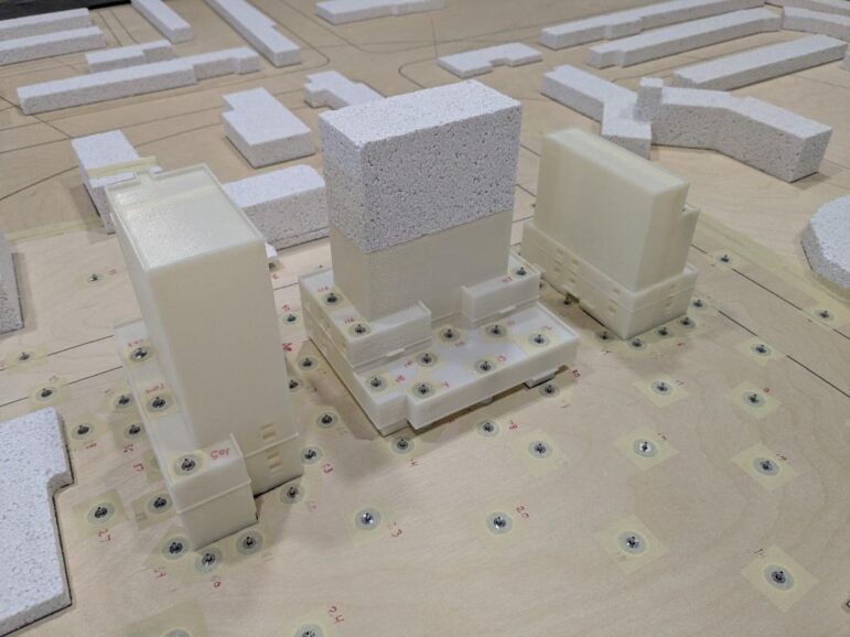A close-up photograph of three white 3D-printed architectural models representing high-rise buildings. They are mounted on a tan-coloured board and surrounded by dozens of small, numbered circular sensors embedded in the surface. In the background, simplified grey foam blocks represent the surrounding neighborhood.