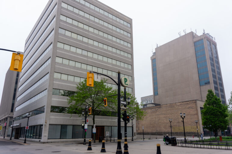 A wide shot of a downtown street corner showing a 10-storey grey concrete building with horizontal bands of windows. Traffic lights and a green "P" parking sign are visible in the foreground. In the background, a large vacant lot is fenced off next to another high-rise building.