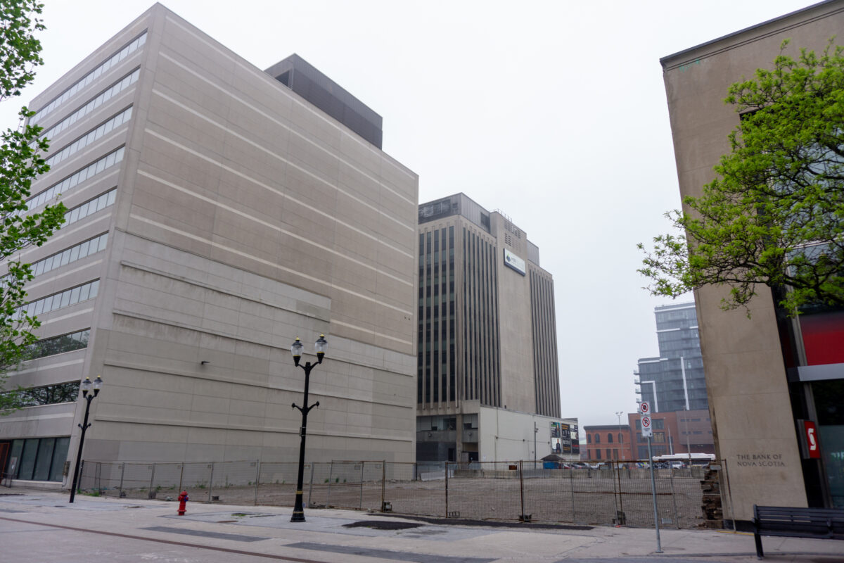 A street-level view looking east along a pedestrian promenade. A large gravel-filled vacant lot is enclosed by a tall chain-link fence, situated between a modern 10-storey concrete building on the left and the stone facade of a Scotiabank building on the right. Modern streetlamps stand along the path under a grey sky.