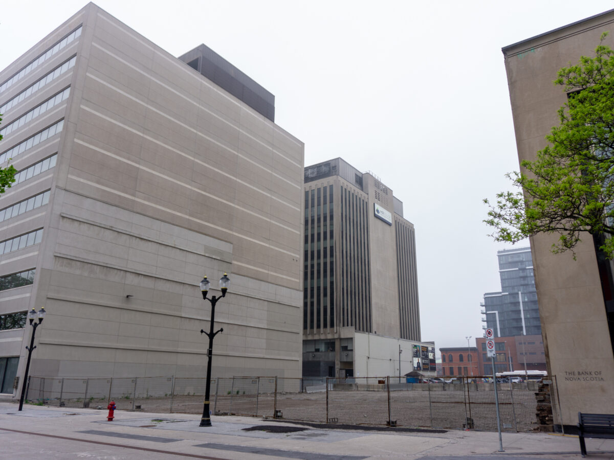 A street-level view looking east along a pedestrian promenade. A large gravel-filled vacant lot is enclosed by a tall chain-link fence, situated between a modern 10-storey concrete building on the left and the stone facade of a Scotiabank building on the right. Modern streetlamps stand along the path under a grey sky.