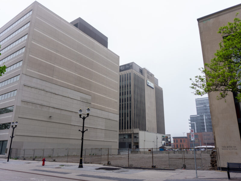 A street-level view looking east along a pedestrian promenade. A large gravel-filled vacant lot is enclosed by a tall chain-link fence, situated between a modern 10-storey concrete building on the left and the stone facade of a Scotiabank building on the right. Modern streetlamps stand along the path under a grey sky.