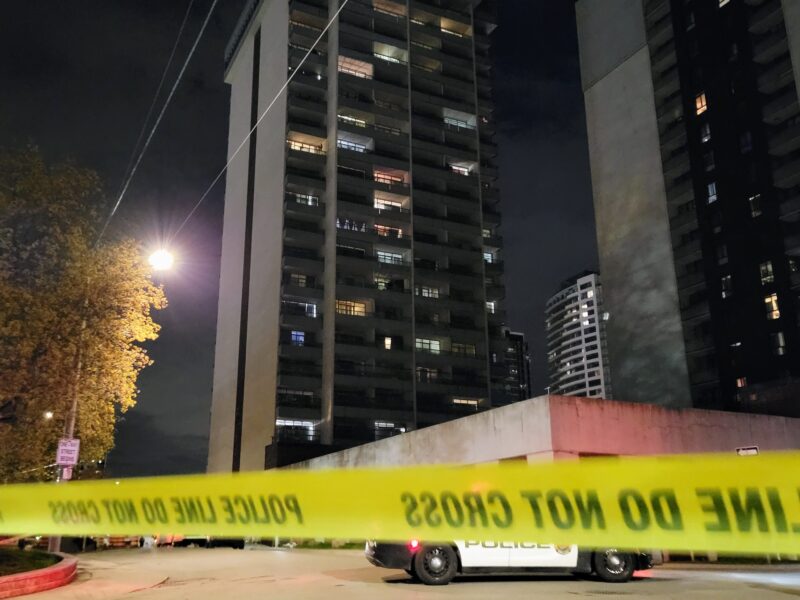 A nighttime view of a high-rise apartment building at 191 Main Street West. In the foreground, yellow "POLICE LINE DO NOT CROSS" tape is stretched across the frame, partially obscuring a parked police cruiser.