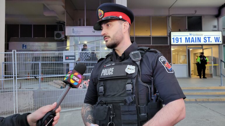 A Hamilton Police officer in a black uniform and duty cap stands in front of the CityHousing Hamilton building entrance labeled "191 MAIN ST. W." He is speaking into a microphone held by a reporter from CHCH News.