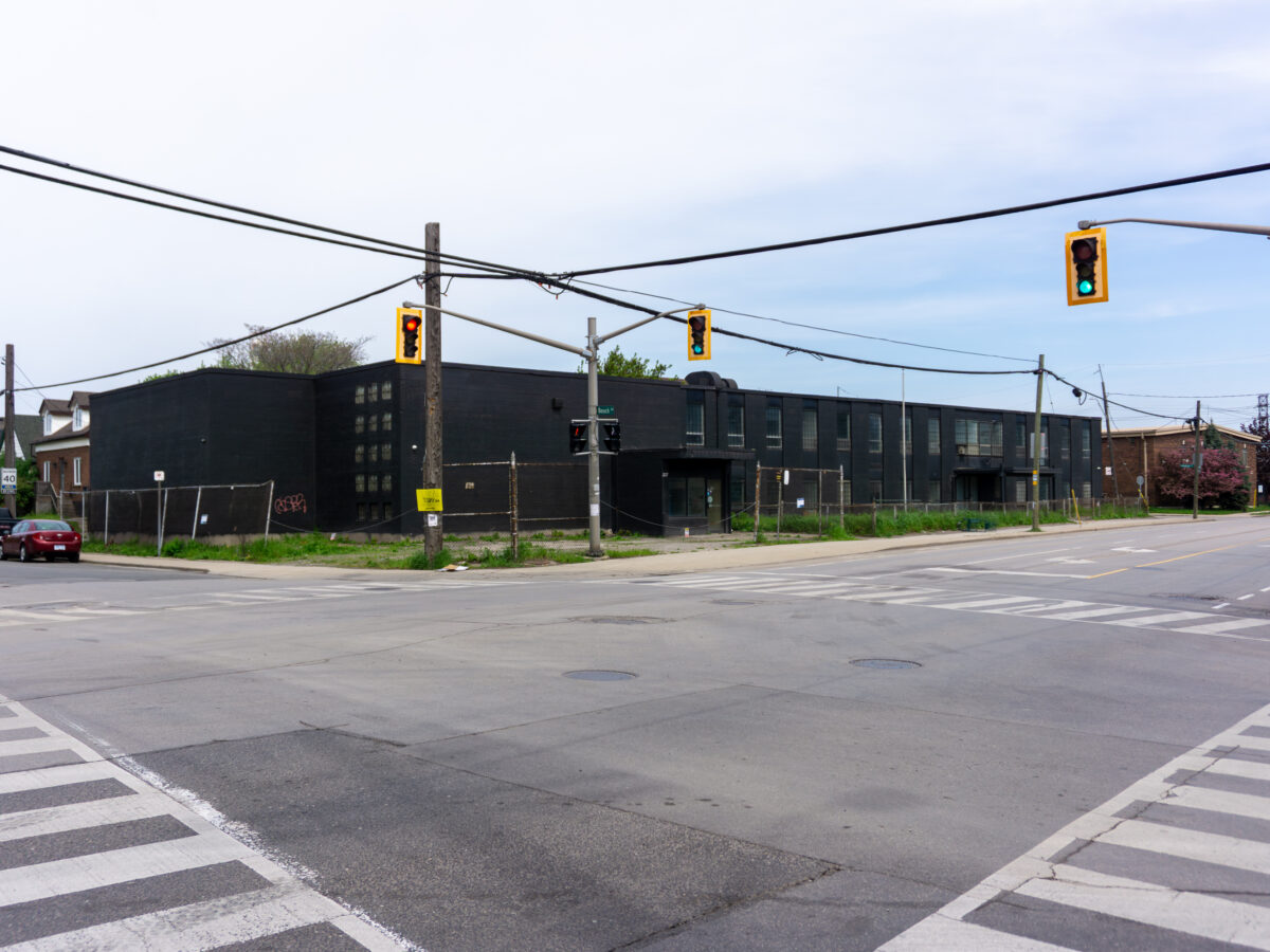 A wide shot looking across a multi-lane street intersection at a large, two-storey commercial building painted entirely matte black. The building features small, square glass-block windows, a chain-link security fence along the property line, and traffic lights hanging over the intersection in the foreground under an overcast sky.
