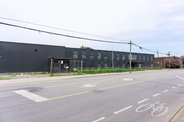 A straight-on shot from across the street showing a long, black-painted brick industrial building. A bicycle lane stencil is visible on the asphalt in the bottom right foreground. The property is enclosed by a chain-link fence with overgrown weeds along the base.