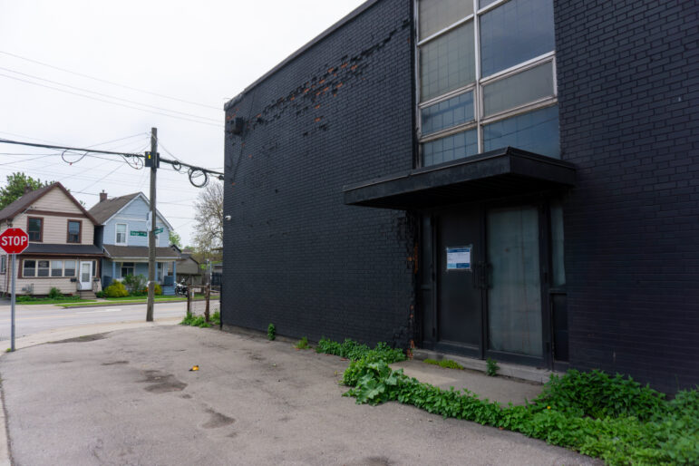 A close-up shot of a recessed double-door entrance with a small overhead awning on a matte black brick wall. To the left, across the street, two residential houses—one beige and one light blue—are visible next to a stop sign at a street corner.