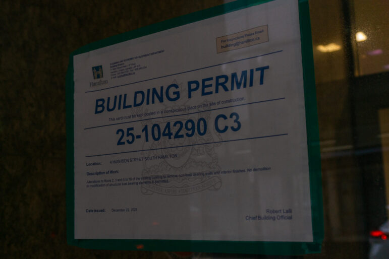 A close-up shot of an official white City of Hamilton building permit taped to a glass window. The permit number "25-104290 C3" is printed in large blue font. The description text notes alterations to floors 2, 3, and 5 to 10 to remove non-load bearing walls and interior finishes, specifying no structural demolition is permitted.
