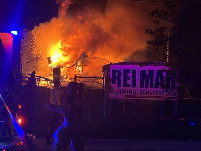 A nighttime dramatic shot showing a massive, bright orange fire engulfing a structure or pile behind a fence. In the foreground, a silhouetted first responder stands near a flashing blue emergency light from a fire apparatus, and a sign reading "REIMAR" is partially illuminated by the flames.
