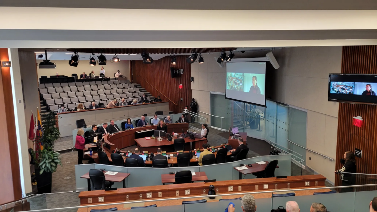 A wide, high-angle interior shot of the Hamilton City Council Chambers. Council members and staff are seated at the large, curved, horseshoe-shaped desk in the center of the room. A large projection screen on the wall shows a video feed of a remote participant. The gallery seating is visible in the background, with several attendees present, and the room is lit by overhead studio lighting.