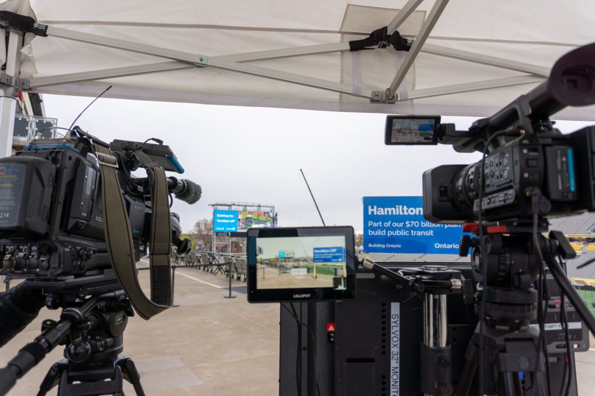 Two professional video cameras on tripods positioned under a white canopy tent at Hamilton Stadium, pointed toward the announcement area. A blue Hamilton LRT banner and a large LED display screen are visible in the background.