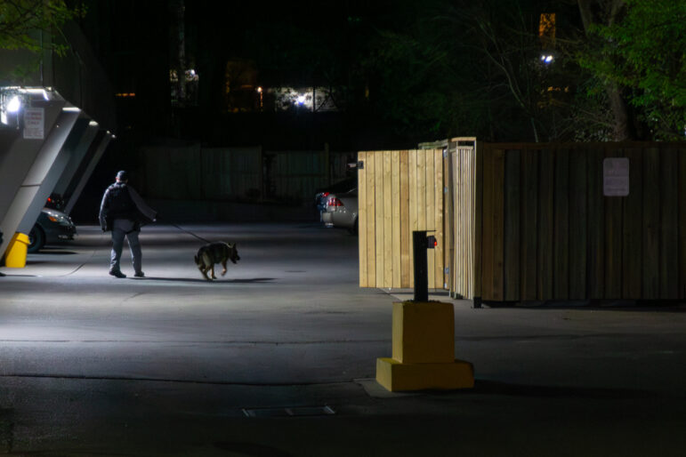 A police officer in dark tactical uniform leads a German Shepherd on a leash through a dimly lit parking lot and alleyway at night, searching near a tall wooden fence.