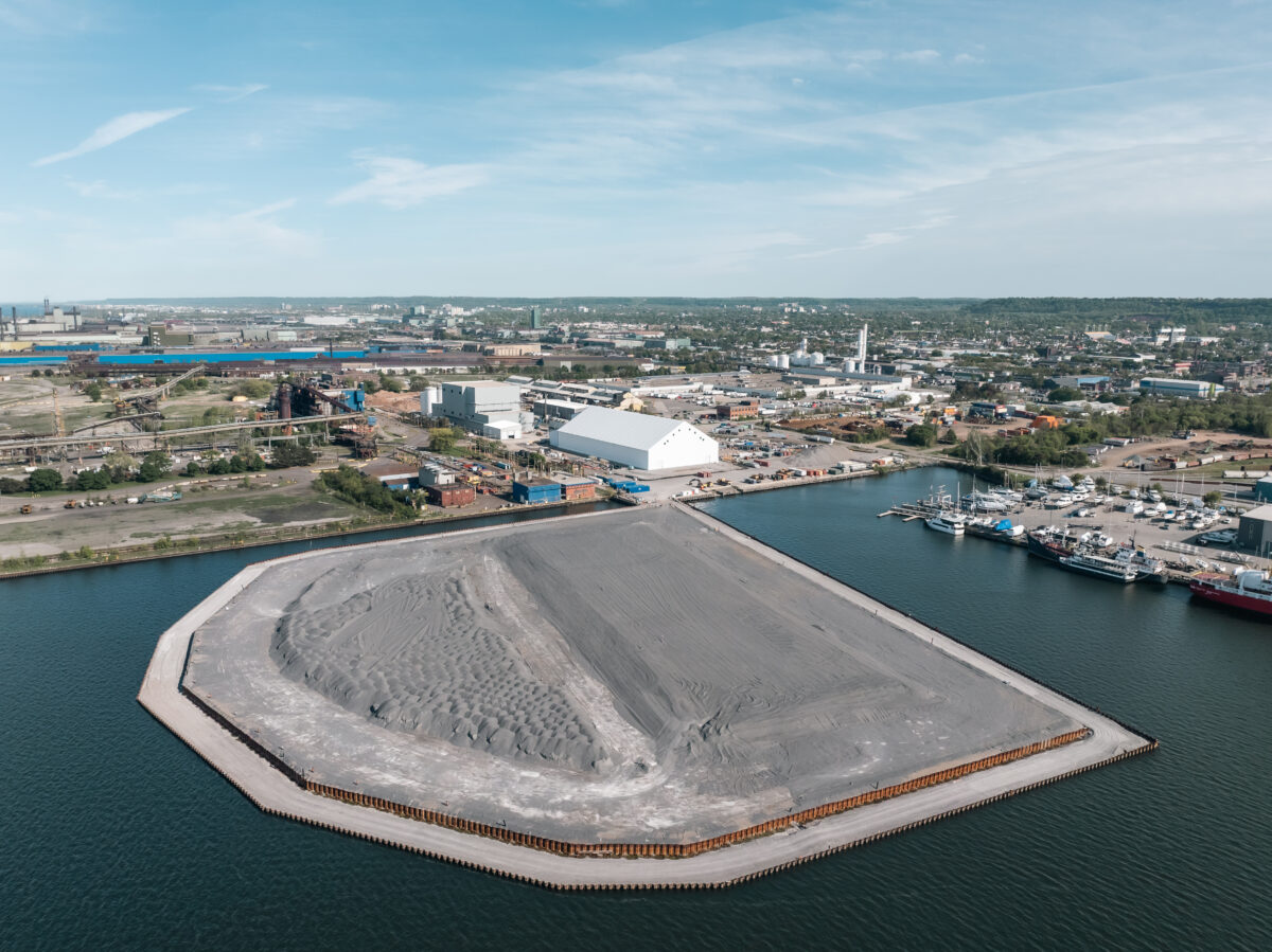 An aerial drone shot looking south across the Hamilton Harbour toward the lower city and the Niagara Escarpment. In the foreground, a massive, shield-shaped engineered landmass enclosed by steel sheet piling sits in the water, surfaced with grey aggregate. Directly behind it on the shore are a large white industrial storage structure and a tall grey processing building, surrounded by various port and manufacturing facilities.