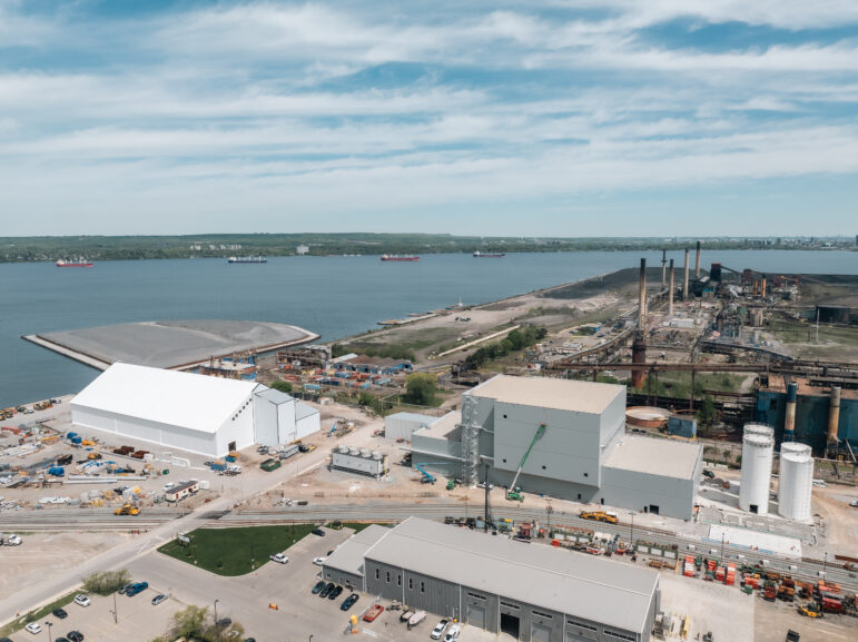 An elevated aerial view looking north over the Hamilton Harbour towards the opposite shoreline. In the foreground center, a large white tent-like storage building and a newly constructed, tall grey industrial facility stand next to railway tracks. To the right, the dark, heavy infrastructure and tall smokestacks of a steel mill are visible. To the left, the edge of the aggregate-topped Randle Reef containment facility juts into the blue water, where several cargo ships are anchored in the distance under a partly cloudy sky.