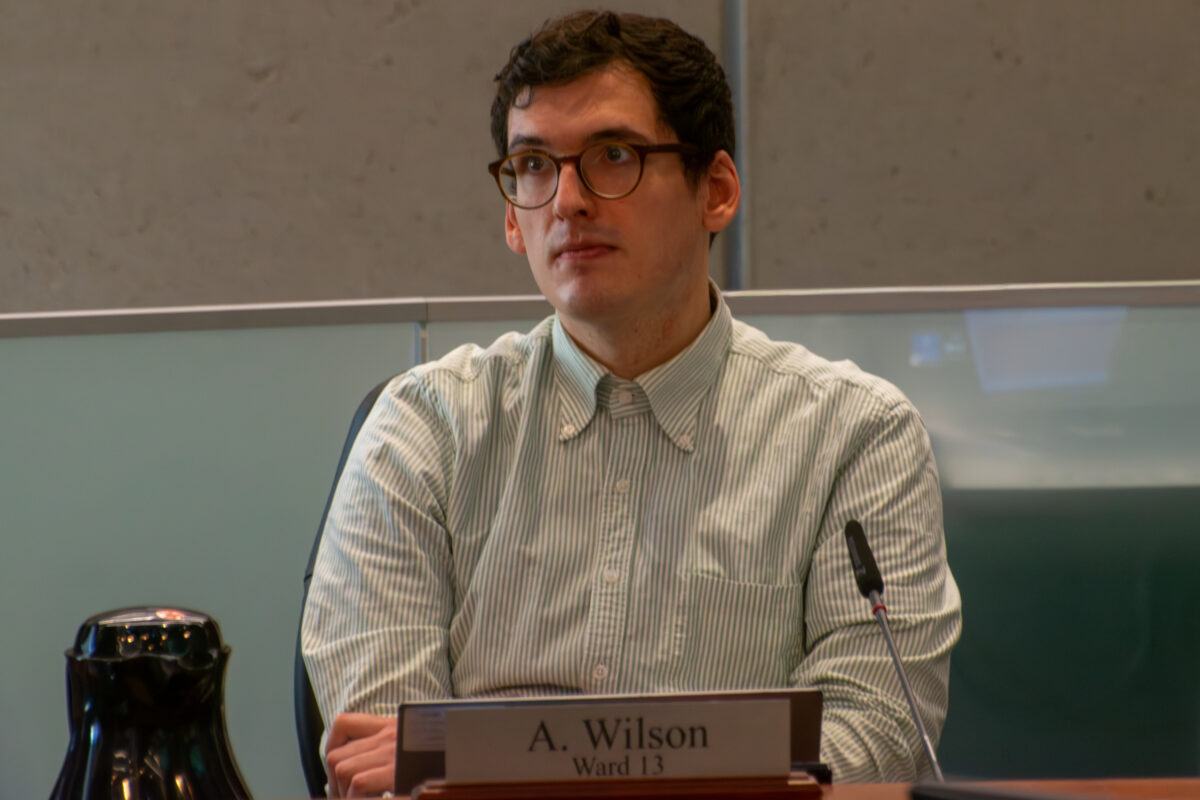 A medium shot of a man with dark wavy hair and glasses, wearing a light green and white pinstriped button-down shirt, seated at a desk in a formal chamber. A nameplate in front of him reads "A. Wilson Ward 13." A black microphone is positioned to his right, and a black water carafe is visible on the left.