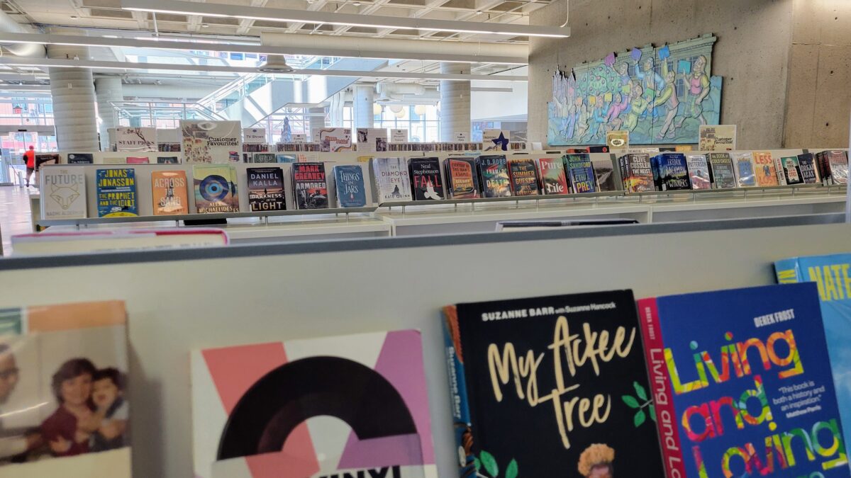 A layered, eye-level shot focuses on rows of hardcover and paperback books arranged face-out on modern display shelves within a bright, open-concept public library. The foreground features close-up, slightly out-of-focus book covers including titles like My Ackee Tree and Living and Loving, while the midground displays a long, well-illuminated shelf holding fiction and non-fiction titles beneath small genre signage placards. The background reveals the expansive, multi-level architectural interior of the facility, characterized by exposed concrete columns, high ceilings, a distant staircase, and a large, colourful artistic mural depicting stylized figures mounted on a concrete wall under cool, even indoor lighting.