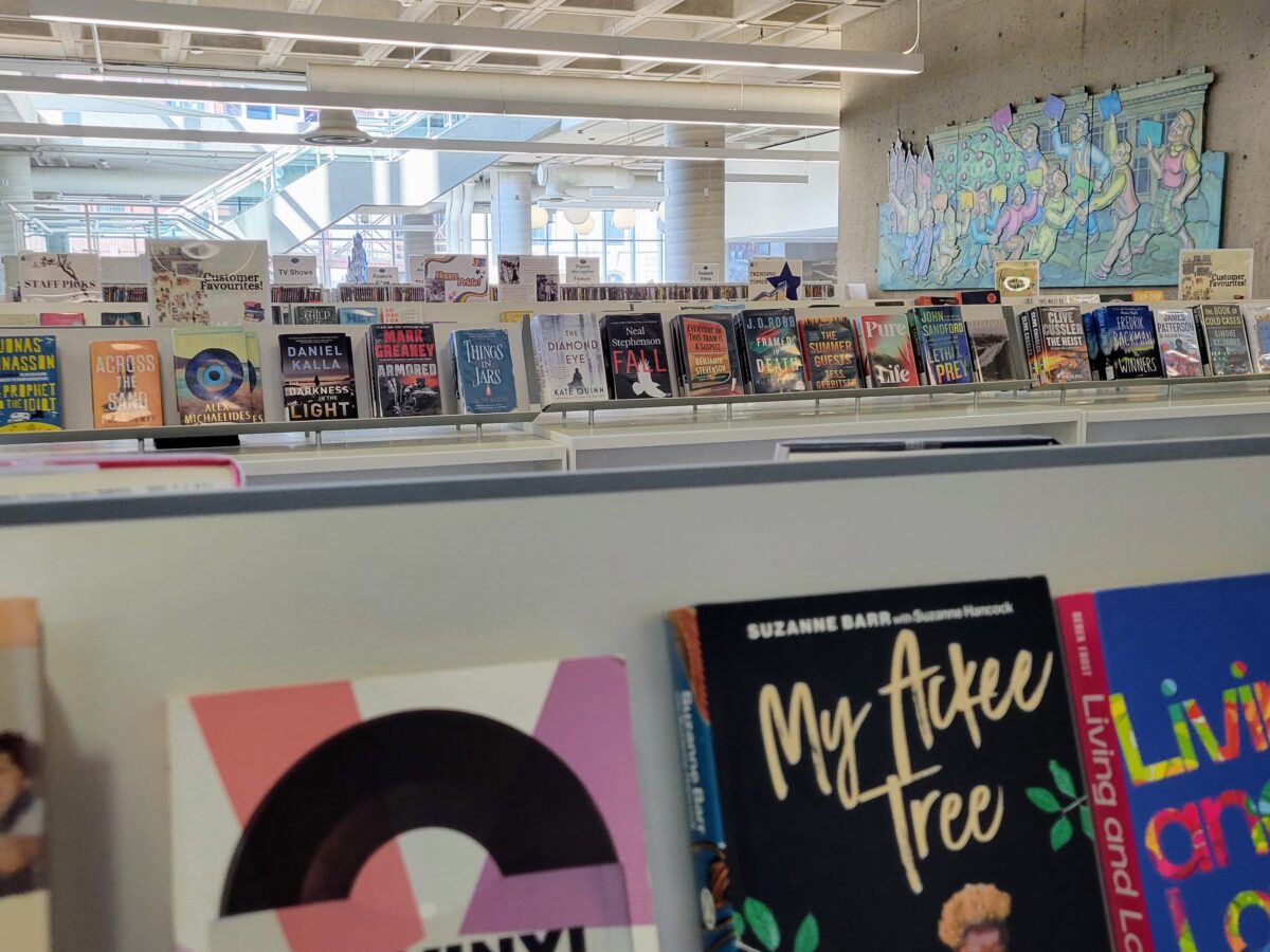 A layered, eye-level shot focuses on rows of hardcover and paperback books arranged face-out on modern display shelves within a bright, open-concept public library. The foreground features close-up, slightly out-of-focus book covers including titles like My Ackee Tree and Living and Loving, while the midground displays a long, well-illuminated shelf holding fiction and non-fiction titles beneath small genre signage placards. The background reveals the expansive, multi-level architectural interior of the facility, characterized by exposed concrete columns, high ceilings, a distant staircase, and a large, colourful artistic mural depicting stylized figures mounted on a concrete wall under cool, even indoor lighting.