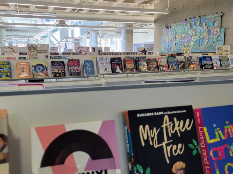 A layered, eye-level shot focuses on rows of hardcover and paperback books arranged face-out on modern display shelves within a bright, open-concept public library. The foreground features close-up, slightly out-of-focus book covers including titles like My Ackee Tree and Living and Loving, while the midground displays a long, well-illuminated shelf holding fiction and non-fiction titles beneath small genre signage placards. The background reveals the expansive, multi-level architectural interior of the facility, characterized by exposed concrete columns, high ceilings, a distant staircase, and a large, colourful artistic mural depicting stylized figures mounted on a concrete wall under cool, even indoor lighting.