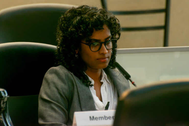 A tight close-up shot focuses on a woman with dark, curly hair and glasses, dressed in a grey blazer over a white collared shirt. She looks downward with a serious expression, seated at a meeting table behind a desk microphone. A small name card reading "Member" is partially visible in the lower foreground, set against a softly blurred background of office chairs and frosted glass privacy panels under balanced indoor lighting.