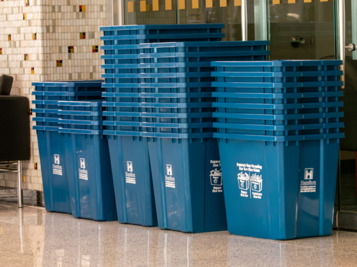 A medium shot of multiple tall stacks of bright blue plastic recycling bins lined up against a wall with small, multi-coloured square mosaic tiles. The bin in the foreground displays white printed graphics reading "Separate Your Recycling - Use Two Blue Boxes" along with the official City of Hamilton logo. The glossy, polished stone floor reflects the base of the bins.