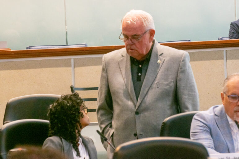 A medium shot documents an interaction inside a civic chamber, featuring a man in a grey blazer standing and looking down toward a seated woman with dark curly hair who is looking back up at him. Another board member in a light blue suit jacket is partially visible in the lower right foreground. The background consists of a frosted glass partition and light wood panelling under clean, bright institutional lighting.