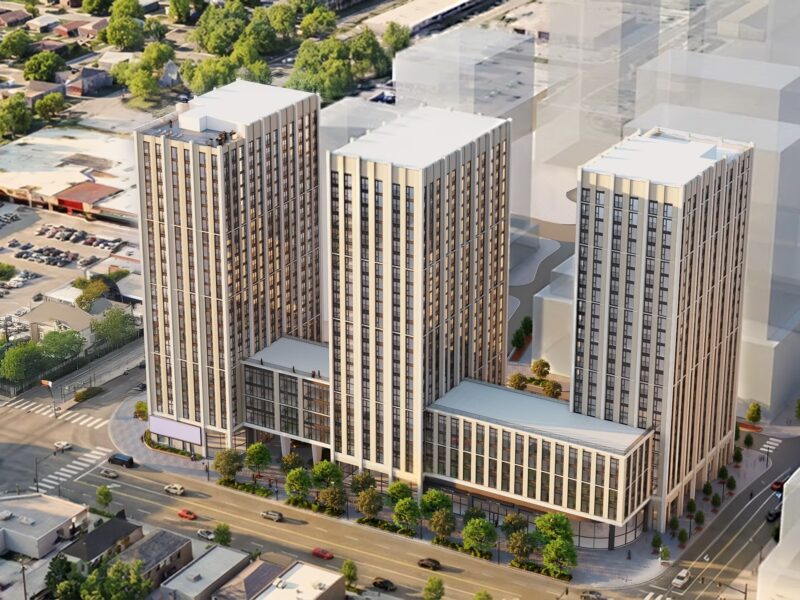 An aerial, bird's-eye architectural rendering displays a proposed mixed-use complex featuring three contemporary high-rise residential towers rising from connected multi-storey podium structures. The buildings exhibit light-coloured stone or precast cladding panels contrasted by vertical strips of dark-framed windows and rectangular balconies, with green rooftop terraces situated on the podium levels. The complex is situated at a busy urban intersection flanked by multiple lanes of vehicle traffic, a suburban commercial plaza parking lot to the left, and low-rise residential neighbourhoods stretching into the distance. Ghosted, semi-transparent grey geometric blocks are positioned to the right to simulate future phase build-outs under clear, artificial daylight conditions designed for urban planning assessment.
