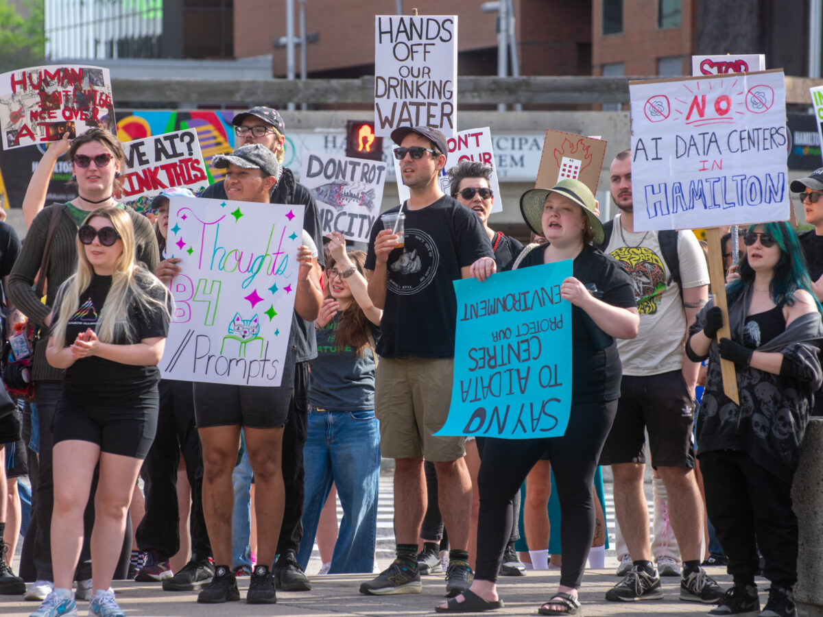 A medium shot of a group of demonstrators holding various handmade placards. Notable signs read "HANDS OFF OUR DRINKING WATER," "NO AI DATA CENTERS IN HAMILTON," and "SAY NO TO AI DATA CENTRES | PROTECT OUR ENVIRONMENT." The individuals are wearing casual summer attire and sunglasses.
