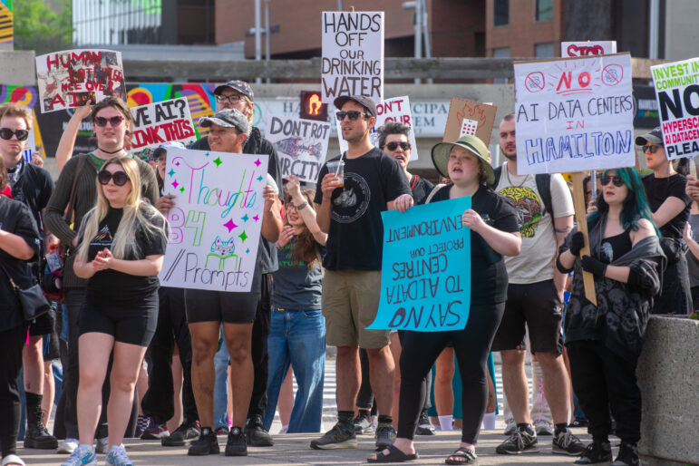 A medium shot of a group of demonstrators holding various handmade placards. Notable signs read "HANDS OFF OUR DRINKING WATER," "NO AI DATA CENTERS IN HAMILTON," and "SAY NO TO AI DATA CENTRES | PROTECT OUR ENVIRONMENT." The individuals are wearing casual summer attire and sunglasses.