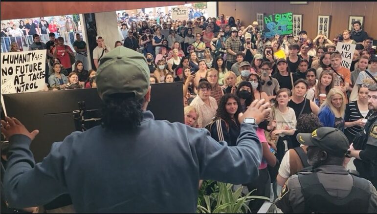 A high-angle, behind-the-shoulder perspective looks out from a podium towards a densely packed crowd filling an indoor public lobby. In the foreground, the back of a person wearing a dark teal zip-up jacket and a green baseball cap is visible with arms slightly outstretched as if addressing the assembly, while a security guard stands to the lower right. The diverse crowd features individuals of varying ages observing the speaker, with several people holding hand-painted protest signs with slogans including "Humanity Has No Future If AI Takes Control" and "No Data Centre". The municipal building interior is illuminated by bright overhead lights, revealing wood-panelled walls, framed portraits, and an upper mezzanine packed with additional observers in the background.