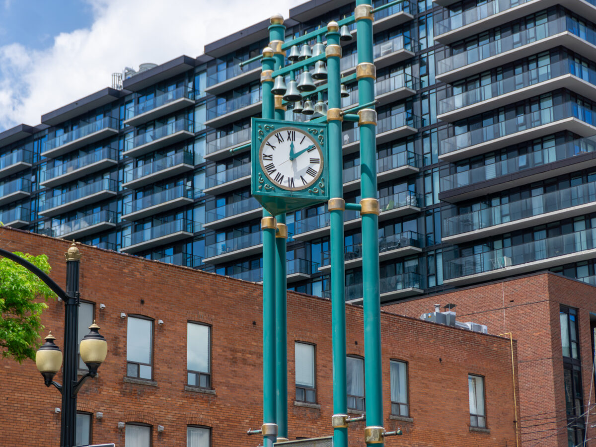 A low-angle, daytime shot captures a teal-coloured public clock tower structure featuring a white clock face with Roman numerals and a small carillon of bells mounted at the top. In the immediate background, a multi-storey heritage red-brick building with regular rectangular windows stretches across the lower half of the frame. Looming behind the brick structure is a contemporary, multi-storey glass and dark-framed residential condominium building with numerous balconies. Bright, natural midday sunlight illuminates the scene under a blue sky scattered with white clouds, with a classic street lamp post visible on the left side.