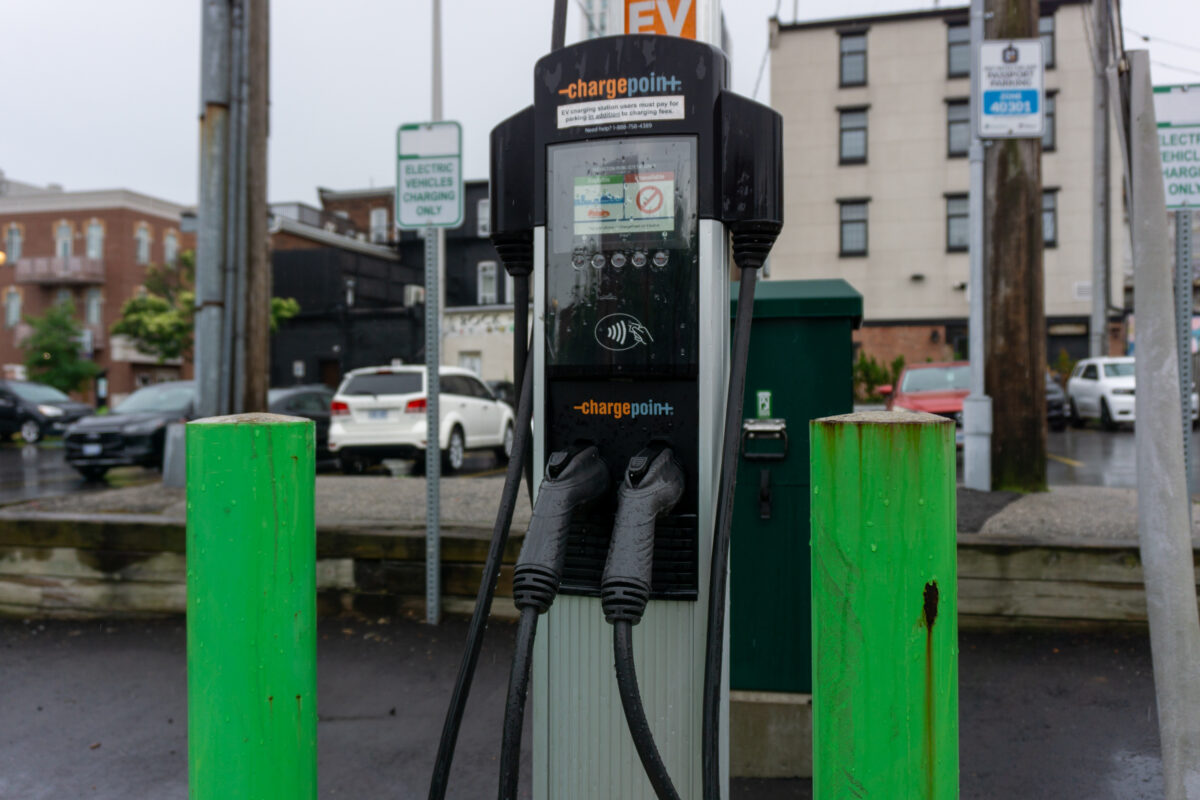 A close-up shot of an outdoor ChargePoint electronic vehicle charging stand flanked by two bright green protective steel bollards. In the background, a rainy asphalt parking lot contains several parked cars and brick residential buildings.