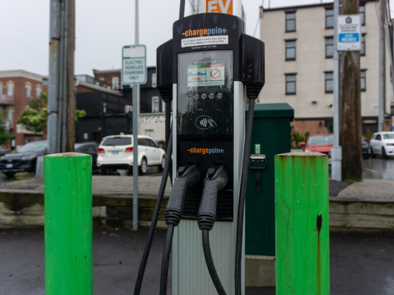 A close-up shot of an outdoor ChargePoint electronic vehicle charging stand flanked by two bright green protective steel bollards. In the background, a rainy asphalt parking lot contains several parked cars and brick residential buildings.