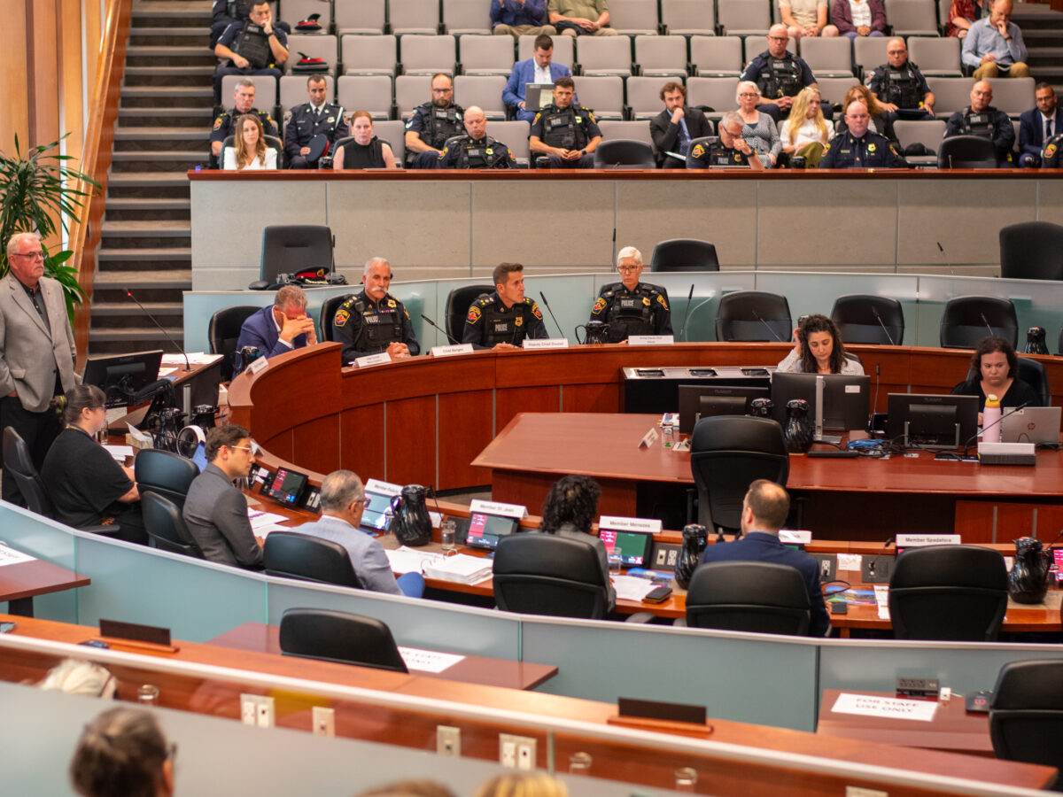A wide, high-angle interior photograph shows a formal meeting underway inside a large, tiered municipal auditorium. Members of the board are seated along a curved wooden dais in the midground, accompanied by uniformed high-ranking police officers. In the foreground, additional staff members sit at long desks equipped with laptops and microphones. The background features steep, stadium-style seating populated by several uniform-clad police personnel and civilian observers beneath modern recessed lighting.